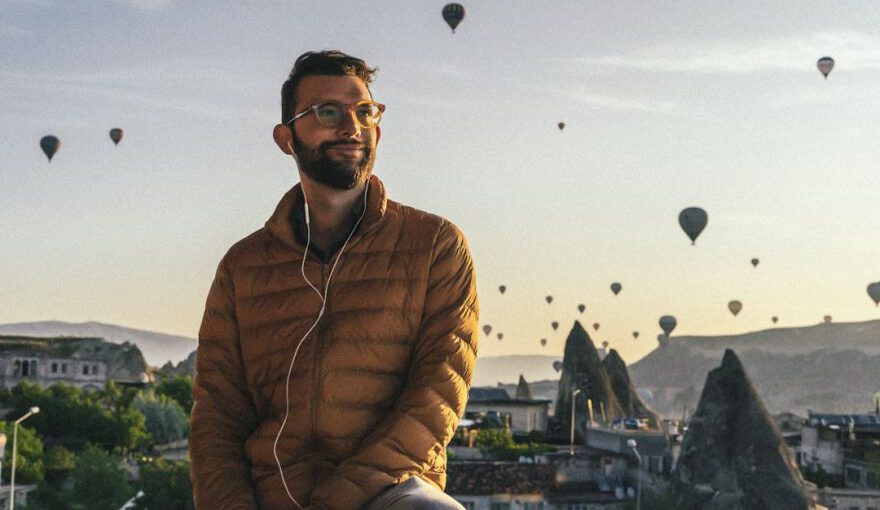 Thermal Relaxation Cappadocia - a man sitting on a ledge with many hot air balloons in the sky