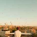 Göreme - a person standing on a balcony looking at hot air balloons in the sky