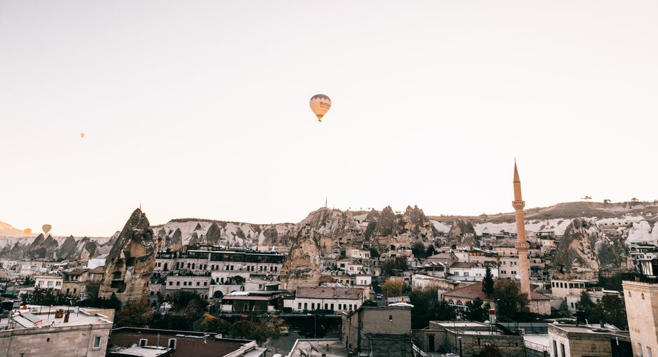 Picturesque scenery of old town with mosque placed among rocky formations in Cappadocia under flying hot air balloon in cloudless sky in daytime